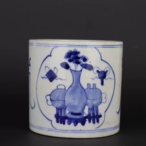 Può includere: Un vaso cilindrico in ceramica bianca con disegni floreali e geometrici blu. Il vaso presenta un disegno centrale di un vaso con fiori, affiancato da due oggetti arrotondati. Il disegno è incorniciato da un bordo blu.