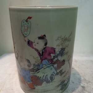 May include: A white ceramic cylinder vase with a hand-painted design of two children in traditional Chinese clothing. The vase features a light green background with bamboo stalks and flowers. The children are holding a lantern with the Chinese character for "good fortune" written on it.