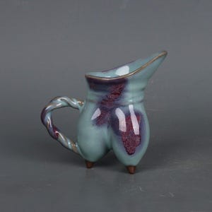May include: A small, ceramic pitcher with a unique design. The pitcher is primarily teal with dark purple accents and a twisted handle. It has three small feet and a gold-colored rim.