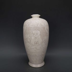 May include: A cream-colored ceramic vase with a detailed etched design. The vase features a central image of a seated figure surrounded by lotus flowers and intricate patterns. The vase has a rounded body and a narrow neck.