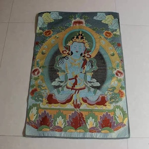 May include: A blue deity with a crown sits in a lotus position on a throne surrounded by a mandala of flowers and gold accents. The deity has a serene expression and holds their hands in a gesture of prayer. The background is a dark blue with clouds and gold accents.