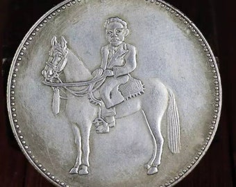Könnte beinhalten: Eine Silbermünze mit einem detaillierten Bild einer Person, die auf einem Pferd reitet. Die Münze hat einen perlenbesetzten Rand.