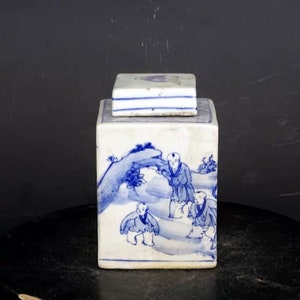 Könnte beinhalten: Eine quadratische Porzellan-Dose mit Deckel, verziert mit einem blauen und weißen Design von Figuren in einer Landschaft. Die Dose ist etwa 13 cm hoch.