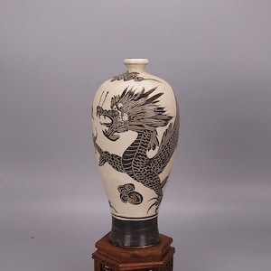 May include: A cream-colored ceramic vase with a black dragon design. The vase has a dark base and a narrow neck. The dragon is depicted with intricate details, and the vase is displayed on a wooden stand.