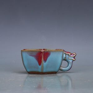May include: A turquoise ceramic cup with a dragon-shaped handle. The cup has a square shape with a red and brown interior. The handle is a light purple color. The cup is likely for tea or coffee.