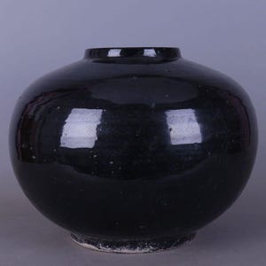 Könnte beinhalten: Eine glänzende, schwarze Keramikvase mit rundem Körper und schmalem Hals. Die Vase hat eine glatte, reflektierende Oberfläche, die das Licht einfängt und Glanzlichter erzeugt. Der Boden der Vase ist etwas breiter als der Hals und steht auf einem kleinen, unglasierten Fuß.