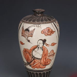 May include: A tall, ceramic vase with a white base and a dark brown top and bottom. The vase features a hand-painted design of a crane, clouds, and a seated figure in shades of brown and red.