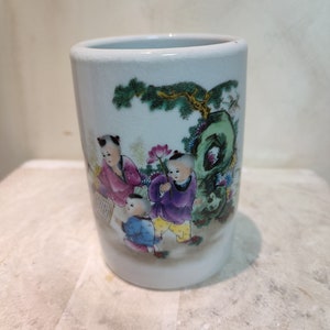 May include: A white ceramic cylinder with a painted scene of children playing in a garden. The children are wearing traditional Chinese clothing and are surrounded by trees, flowers, and a rock formation.