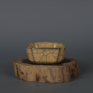 May include: A small, square, ceramic bowl with a crackled glaze in shades of cream and black. The bowl is sitting on a wooden stand.