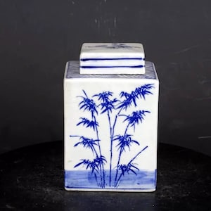 Pode incluir: Um pote quadrado de porcelana branca com um desenho de bambu azul e branco. A tampa também é quadrada e tem um desenho azul e branco.