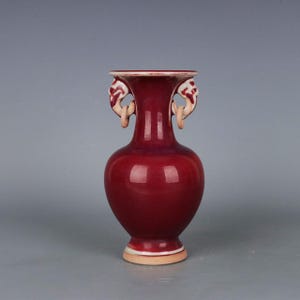 May include: A maroon-colored ceramic vase with a bulbous body and a flared rim. The vase features two decorative handles and a cream-colored base. The vase is a decorative item.