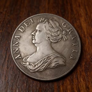 以下が含まれることがあります： 女性の横顔が描かれた銀貨で、「ANNA DEI GRATIA」の文字が刻まれています。コインは精巧なデザインで、少し使い込まれた外観をしており、古さを感じさせます。コインは丸く、縁はざらざらしています。