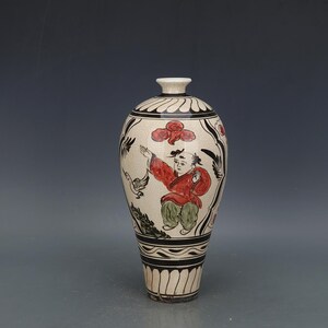 May include: A decorative ceramic vase with a cream-colored background and black and red painted details. The vase features a design of a person in a red coat and green pants, with black floral and geometric patterns. The vase has a narrow neck and a wide base.