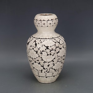Könnte beinhalten: Eine weiße Keramikvase mit schwarzen Blumen- und Vogelmustern. Die Vase hat einen bauchigen Körper und einen schmalen Hals mit dekorativem Rand. Die aufwendigen Muster bedecken die gesamte Oberfläche und erzeugen einen auffälligen Kontrast.