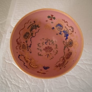 Puede incluir: Un cuenco de cerámica rosa con un diseño dorado y azul que presenta animales estilizados y motivos florales. El cuenco tiene un borde ligeramente elevado.