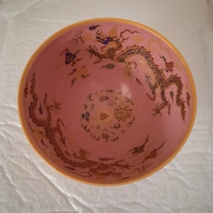 Peut inclure: Un bol en céramique rose avec un motif de dragon doré. Le dragon est enroulé autour du bord du bol et a des accents bleus et dorés. Le bol a un bord doré et un fond blanc.