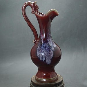 May include: A maroon ceramic pitcher with a curved handle and a flared spout. The pitcher has a bulbous body and a dark blue and white pattern. It sits on a wooden stand, showcasing a classic design.