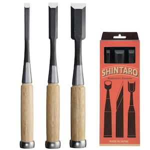 May include: Three wood chisels with black blades and light wood handles are displayed next to a red box. The box features the text "SHINTARO Authentic Chisels" and illustrations of the tools. The chisels have silver ferrules and are arranged vertically.