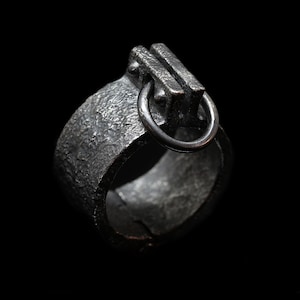 May include: A silver ring with a textured band and a unique, industrial-style design. The ring features a rectangular top with a small, circular loop.