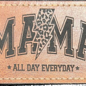 May include: A rectangular brown leather patch with the word "MAMA" in bold black letters. A lightning bolt with a leopard print design is in the center. The text "ALL DAY EVERYDAY" is below the word "MAMA".