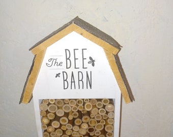 May include: A white wooden bee barn with a brown roof and a rectangular opening filled with bamboo tubes. The text "The BEE BARN" is printed on the front, along with bee illustrations. The barn is designed to attract bees.