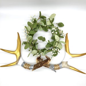 May include: A decorative arrangement featuring a green and white wreath, two gold-painted antlers, and two antler bases with silver and gold accents. The items are arranged on a white background, creating a rustic, natural aesthetic.