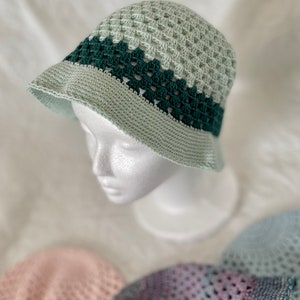 Op de afbeelding: Een groene en lichtgroene gebreide bucket hat met een brede rand. De hoed zit op een wit mannequin hoofd.