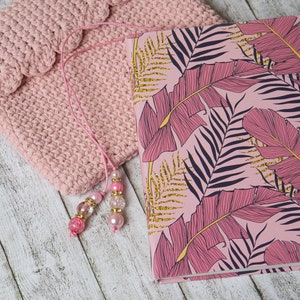 Può includere: Una copertina per libro in crochet rosa con un diario con motivo a foglie tropicali rosa e dorate e un segnalibro con perline rosa.