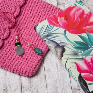 Könnte beinhalten: Eine rosa gehäkelte Tasche mit einem gewellten Rand und einem Perlenquaste. Die Tasche liegt auf einer weißen Holzoberfläche neben einem Buch mit Blumenmuster.