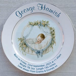 May include: A white ceramic plate with a gold rim, featuring a baby sleeping in a cot surrounded by flowers. The plate is personalised with the name "George Hamish" and the text "Born on Friday, 23rd September, 2023 at 11.40am Weight 7lb 3oz Length 19 1/2 inches Love always from Granny & Grumpy xx".