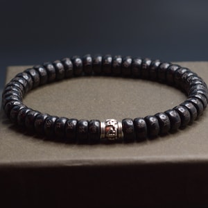 May include: A black wooden beaded bracelet with a silver charm. The bracelet is on a brown box.