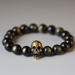 May include: A beaded bracelet featuring black and gold-flecked beads. The focal point is a gold-colored skull charm. The bracelet is likely made of natural stone beads and is designed to be worn around the wrist.