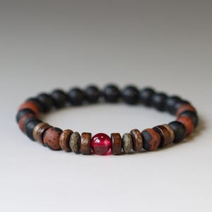 May include: A beaded bracelet featuring a central glossy red spherical bead flanked by dark brown wooden discs, textured grey stone spacers, and matte black spherical beads that continue around the entire circumference of the circular accessory.