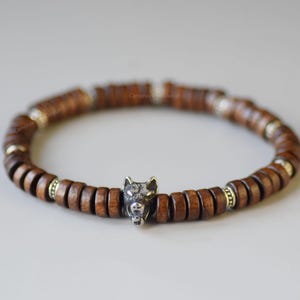 Puede incluir: Una pulsera de cuentas de madera marrón con un dije de cabeza de lobo plateado. La pulsera presenta cuentas redondas de madera marrón oscuro y cuentas plateadas. El dije de cabeza de lobo está centrado.