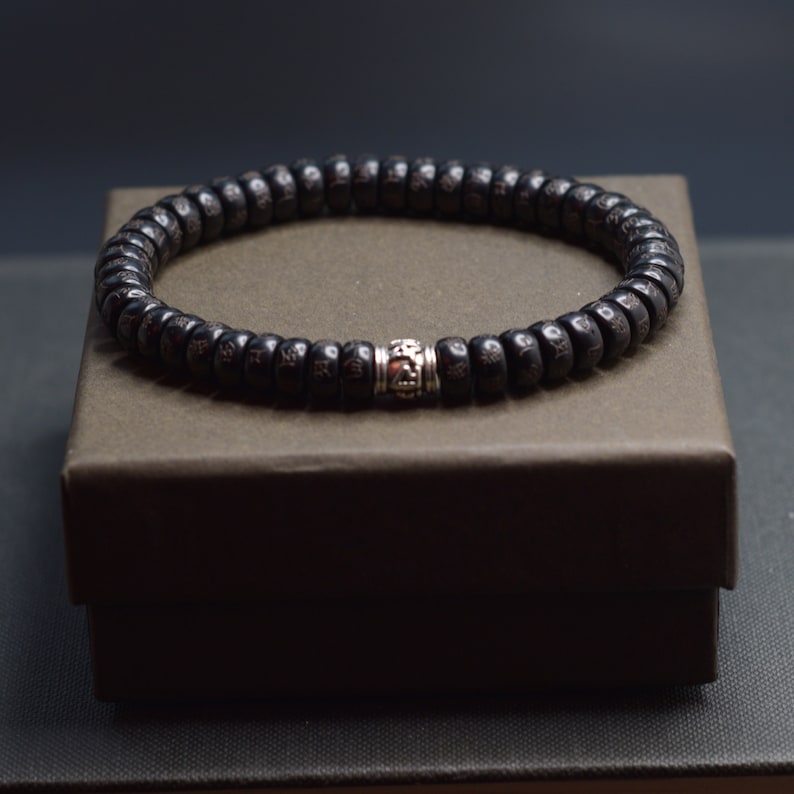 May include: A black wooden beaded bracelet with a silver charm. The bracelet is on a brown box.