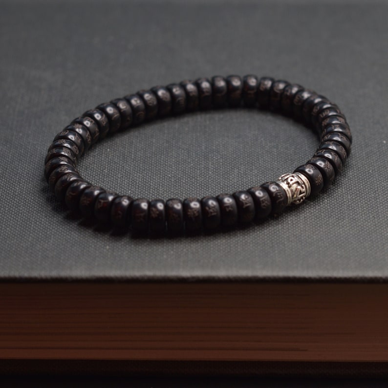May include: A black wooden beaded bracelet with a silver charm. The bracelet is made of small, round beads and has a single silver charm in the center.