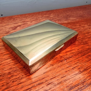 May include: A rectangular, gold-toned metal box with a textured lid. The box has a ridged edge and a subtle wave pattern on the top surface. It rests on a reddish-brown wooden surface.