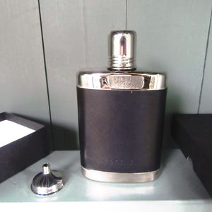 Könnte beinhalten: Eine schwarze Flasche mit silbernem Verschluss und Zierleisten, einem kleinen silbernen Trichter und einer schwarzen Box. Die Flasche hat eine strukturierte Oberfläche und eine rechteckige Form. Die Flasche ist etwa 13 cm hoch.