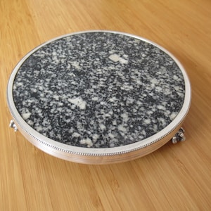 May include: A circular serving tray featuring a speckled black and white granite inlay set within a polished silver-toned metal frame. The tray is supported by three small, decorative metal feet and has a beaded edge along the rim.
