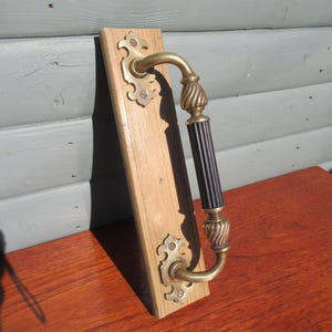 May include: Antique door pull with a brass handle and decorative accents. The handle has a black center section and is mounted on a wooden board. The brass hardware has an ornate design, adding a vintage aesthetic.