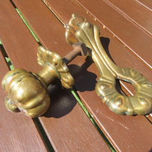 Peut inclure: Deux poignées de porte anciennes en laiton avec des motifs ornés inspirés de fleurs. L'une est un bouton, l'autre une poignée. Le laiton a une patine vieillie, suggérant l'âge. Les poignées reposent sur une surface en bois brun.