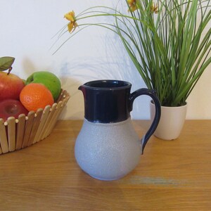 Op de afbeelding: Een grijze en blauwe keramische kan met een getextureerd oppervlak. De kan heeft een brede basis en een smalle hals. De kan staat op een houten tafel.