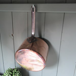 May include: A copper saucepan with a long handle, hanging against a gray wooden wall. The pot has a rounded base and a shiny, reflective surface. The handle is attached with rivets, and the pot appears to be vintage.