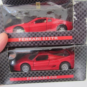 May include: Two red Ferrari model cars in their original boxes. The top box is labeled "FERRARI 512TR" and the bottom box is labeled "FERRARI F50". The boxes have a black and gray checkered pattern.