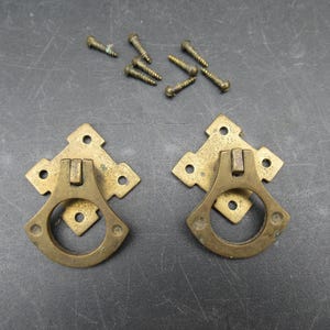 May include: Two antique brass drawer pulls with ring handles and mounting screws. The pulls have a diamond-shaped backplate with a decorative design and a circular handle. The screws are small and brass-colored.