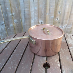 May include: A copper saucepan with a lid and a long handle. The pot is round with a shiny finish. The handle and lid handle are gold-coloured. The pot is sitting on a wooden table.