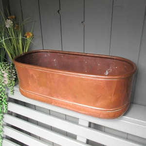 Op de afbeelding: Een langwerpige, ovaalvormige koperen plantenbak met een gladde, metalen afwerking en een afgeronde rand. De bak heeft een diepe, holle binnenkant en een stevige bodem, die de natuurlijke roodbruine tinten van het koperen materiaal laat zien.