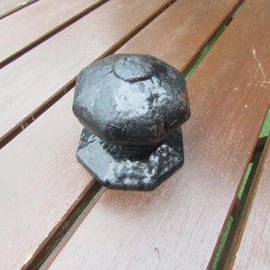 Op de afbeelding: Een zwarte deurknop in antieke stijl met een facetgeslepen, afgeronde bovenkant en een zeshoekige basis. De knop heeft een getextureerd, verouderd uiterlijk, wat suggereert dat hij van gietijzer of een vergelijkbaar materiaal is gemaakt. Hij ligt op een houten oppervlak.