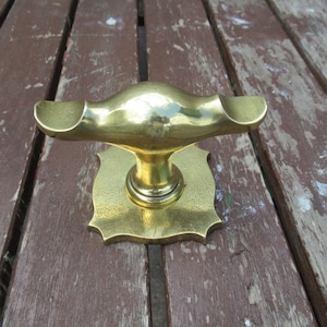 Könnte beinhalten: Ein polierter Türgriff aus Messing mit einem horizontalen T-förmigen Griff mit geschwungenen Enden und einer quadratischen Grundplatte mit gewellten Kanten. Die Metalloberfläche hat ein reflektierendes, glattes Finish mit einem zentralen Pfosten.