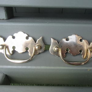 Op de afbeelding: Twee antieke messing lade-grepen met een sierlijk ontwerp. Elke greep heeft een gebogen handvat en een achterplaat met een gestileerde, wolkvormige vorm. De grepen staan tegen een gedempte grijze achtergrond.